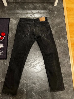 Svarta jeans Jack and Jones - Snygga svarta Jack and Jones jeans.Jeansen har fem fickor, knappgylf och en läderpatch bak i midjan. Tillverkade i slitstarkt denimtyg som ger en tidlös look. Perfekta för dig som gillar en enkel och stilren stil. Strl 30 32