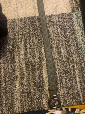 Gucci svart bälte med GG-spänne - Snyggt svart bälte från Gucci med klassiskt GG-mönster längs hela remmen och ett stort, silverfärgat GG-spänne. Bältet har en stilren design och är tillverkat i skinn med detaljer i silvermetall. Perfekt för att lyfta vilken outfit som helst.