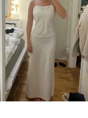 Vit långklänning med tunna band - Elegant vit långklänning med tunna axelband och rak passform. Klänningen har en enkel och stilren design med lätt glansigt material som ger en exklusiv känsla. Perfekt för sommarens speciella tillfällen.