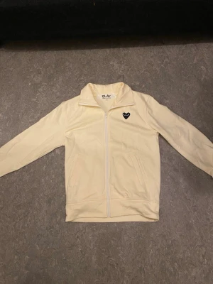 Beige ziptröja Comme des Garçons Play - Snygg beige ziptröja från Comme des Garçons Play med svart hjärta och ögon på bröstet. Tröjan har hög krage, dragkedja framtill och två fickor. Tillverkad i mjukt bomullsmaterial som känns skönt mot huden.