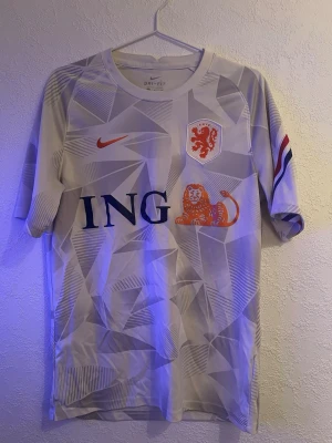 Holland Nike fotbollströja vit - Snygg vit fotbollströja från Nike med Nederländernas landslagsmärke och ING-logga på bröstet. Tröjan har geometriskt mönster, orange lejon och röd-blå-vit detalj på ärmen. Tillverkad i lätt och ventilerande Dri-FIT-material, perfekt för träning eller match.