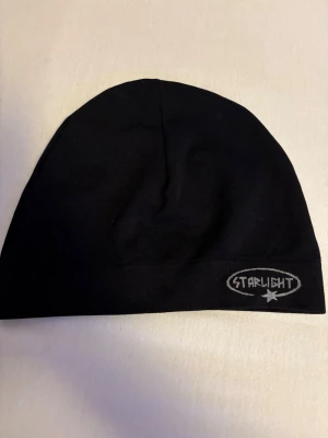 Svart mössa från Starlight - Svart, enkel och stilren mössa från Starlight med broderad logga framtill. Mössan är tillverkad i mjukt tyg och har en klassisk, rundad form som sitter skönt på huvudet. Perfekt accessoar för kyliga dagar.