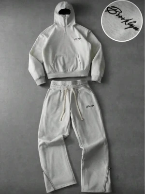 Grått Brooklyn loungewear-set - Snyggt grått loungewear-set med hoodie och byxor från Brooklyn. Hoodien har dragkedja vid halsen och broderad logga på bröstet. Byxorna har resår i midjan med snörning, raka ben och matchande logga. Perfekt för chill dagar hemma.