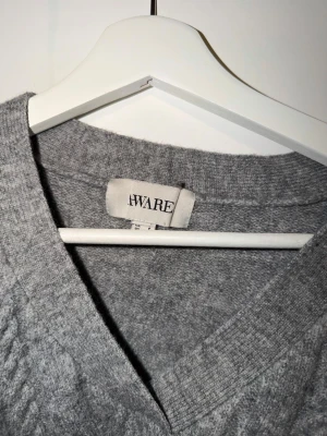 Grå kabelstickad tröja från Aware - Mysig grå kabelstickad tröja från Aware med V-ringning och avslappnad passform. Tröjan har ribbade muddar och ett klassiskt stickat mönster som ger en tidlös look. Perfekt att bära över en t-shirt eller skjorta för extra lager.