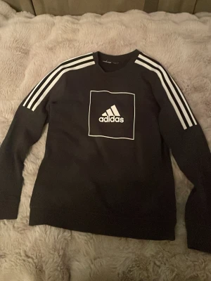 Svart Adidas sweatshirt med vita ränder - Svart sweatshirt från Adidas med klassiska vita ränder längs axlarna och ett vitt Adidas-logotyptryck på bröstet. Tröjan har rund halsringning och är tillverkad i mjukt material, perfekt för en sportig och avslappnad stil. (Barnstolek)