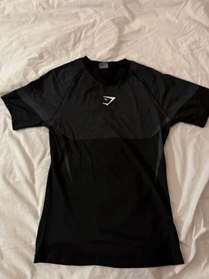 Svart Gymshark T-shirt - Snygg svart tränings tshirt från Gymshark i storlek S med mönstrad ovandel. Inprincip nyskick, endast testad fåtal gånger. T-shirten har en vit Gymshark-logga på bröstet och är tillverkad i ett lätt och andningsbart material, perfekt för gymmet eller löprundan.