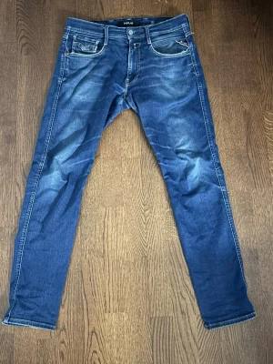 Blå slim fit jeans från Replay - Snygga blå slim fit jeans från Replay med klassisk femficksdesign och slitna detaljer framtill och baktill. Jeansen har normal midja, dragkedjegylf och kontrastsömmar. Tillverkade i denim med lätt stretch för skön passform.