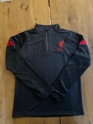 Liverpool FC svart träningsjacka - Svart träningsjacka från Liverpool FC med halv dragkedja och röd broderad logga på bröstet. Ärmarna har diskreta svarta mönster och röda detaljer vid axlarna. Tillverkad i mjukt  syntetmaterial, perfekt för träning eller chill. Storlek L men passar S