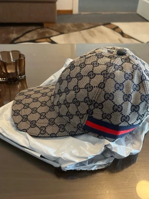 Gucci keps med monogram och röd-blå rand - Säljer en snygg Gucci keps i beige med mörkblått GG-monogram över hela kepsen. Klassisk böjd skärm och justerbar baksida. På sidan finns en ikonisk röd och blå rand. Materialet är canvas med exklusiv känsla.