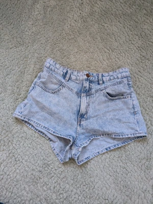Ljusblå denimshorts med hög midja - Snygga ljusblå denimshorts med hög midja och klassisk femficksdesign. Shortsen har bälteshällor, knapp och dragkedja framtill samt råa kanter nedtill för en avslappnad look. Perfekta för varma dagar och enkel att matcha med allt.
