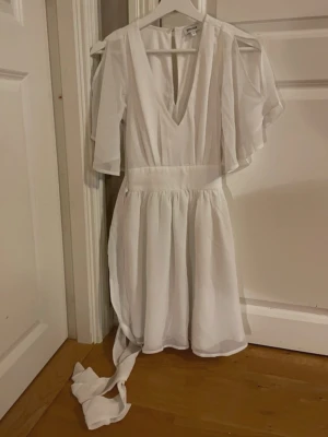 Vit klänning från Bubbleroom - Supersöt vit klänning från Bubbleroom med v-ringning, volangärmar och markerad midja. Klänningen har ett flowigt tyg och ett band i midjan som knyts. Perfekt för sommarens alla tillfällen och ger en fräsch look. Skulle också kunna vara en perfekt studentklänning!