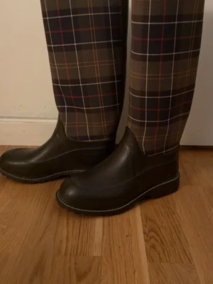 Rutiga gummistövlar med låg klack från barbour - Snygga knähöga gummistövlar med rutigt mönster i brunt, svart, rött och beige från barbour . Stövlarna har en låg klack och rundad tå, perfekta för regniga dagar. Tillverkade i slitstarkt gummi med klassisk design och kontrastsömmar.