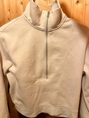 Ljusbeige halfzip hoodie - Ljusbeige hoodie med halv dragkedja och hög krage. Tillverkad i mjukt sweatshirtmaterial med borstad insida för extra komfort. Croppad modell med långa ärmar och stilren design utan tryck eller mönster.