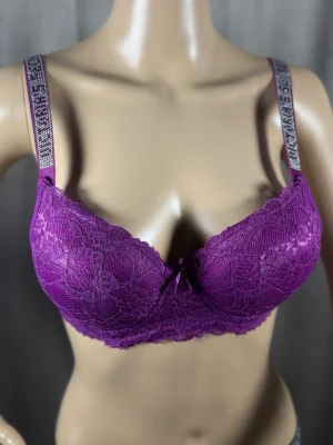 Lila spets-bh med glittriga band - Säljer en snygg lila bh i spets med vadderade kupor och dekorativ rosett framtill. Axelbanden är prydda med glittriga stenar och text, vilket ger en extra trendig touch. Perfekt för dig som gillar detaljer och vill ha något unikt i underklädeslådan.