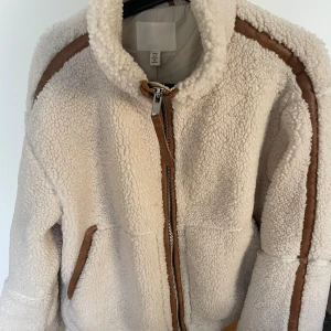 Beige teddyjacka h&m - Mysig teddyjacka från HM i beige med bruna detaljer längs axlar och ärmar. Jackan har hög krage, dragkedja framtill och två fickor. Perfekt för kalla dagar och ger en trendig look med sitt fluffiga material. Lite missfärgad inne men inget som syns utifrån. Kan eventuellt gå bort vid tvätt 