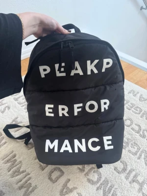 Svart ryggsäck från Peak Performance - Snygg svart ryggsäck från Peak Performance med stora vita bokstäver på framsidan. Ryggsäcken har dragkedja upptill, justerbara axelband och ett vadderat material som ger en cool look. Perfekt för skola eller träning.