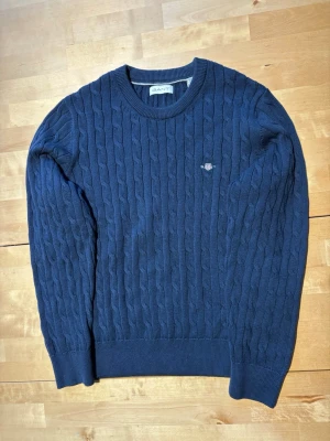 Mörkblå kabelstickad tröja Gant - Kabelstickad mörkblå tröja från Gant med rund halsringning och diskret logga broderad på bröstet. Tröjan har ribbade muddar vid ärmslut och nederkant, vilket ger en klassisk och stilren look. Perfekt för lager-på-lager och enkel att matcha.