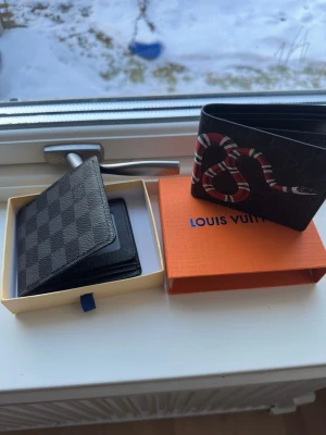 Louis Vuitton och Gucci plånböck - Två snygga plånböcker: en klassisk Louis Vuitton i svart med rutmönster och en Gucci i svart med ett coolt ormmotiv i rött och vitt. Båda är tillverkade i läder och har plats för kort och sedlar. Perfekta accessoarer för dig som gillar lyxiga detaljer.