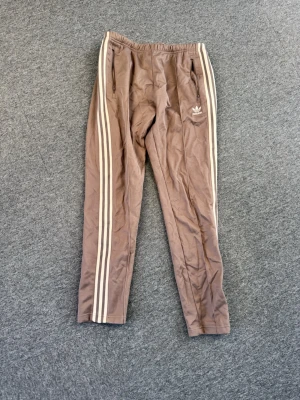 Bruna Adidas mjukisbyxor med ränder - Säljer ett par bruna mjukisbyxor från Adidas med klassiska vita ränder längs benen och Adidas-logga på vänster sida. Byxorna har dragkedjefickor och resår i midjan. Perfekta för chill dagar eller träning. Materialet är mjukt och skönt.