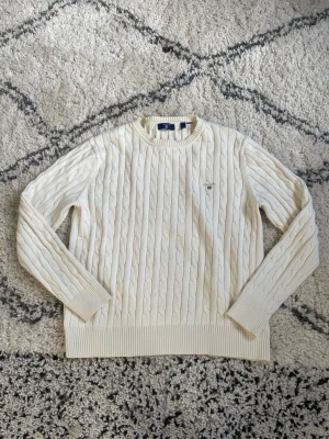 Vit kabelstickad tröja från Gant - Kabelstickad tröja från Gant i mjuk, vit bomull. Klassisk rund halsringning och ribbade muddar vid ärmslut och nederkant. Liten broderad Gant-logga på bröstet. Perfekt för en clean och stilren look.