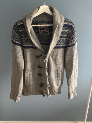 Grå kofta med mönster och träknappar.  - Säljer en grå stickad kofta från Lambretta med mörkblått och vitt nordiskt mönster över axlarna. Koftan har stora träknappar, två fickor framtill och en bred sjalkrage. Perfekt för dig som gillar klassisk och mysig stil.💫 bara höra av dig för mer bilder osv!