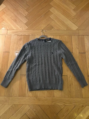 Grå kabelstickad tröja Polo Ralph Lauren - Snygg grå kabelstickad tröja från Polo Ralph Lauren med rund hals och diskret broderad logga på bröstet.  Tröjanr helt ny aldrig använd och har inga defekter alls. Storleken är small hör av vid funderingar!