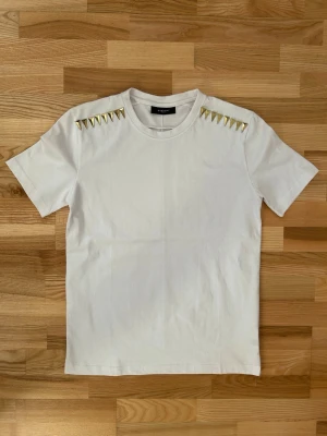 Vit t-shirt med nitar från Givenchy - Säljer en clean vit t-shirt från Givenchy med guldiga pyramidnitar på axlarna som ger en edgy vibe. T-shirten är i 100% bomull, har rund hals och korta ärmar. Perfekt för dig som gillar designerplagg med coola detaljer.
