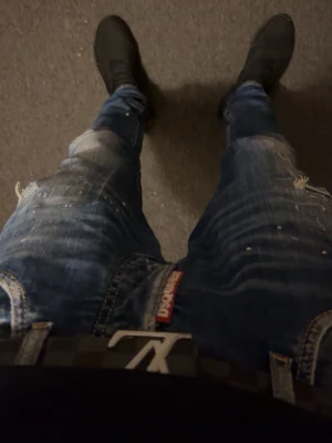 Blå Dsquared2 jeans med slitningar - Säljer ett par blå jeans från Dsquared2 med slitna detaljer och tvättade effekter. Jeansen har en röd Dsquared2-tag vid gylfen och sitter snyggt med en smal passform. Perfekta för dig som gillar en trendig och avslappnad stil.