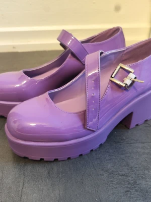 Lila Mary Jane pumps från Koi Footwear - Säljer ett par lila Mary Jane pumps från Koi Footwear med chunky plattform och bred klack. Skorna har en glansig finish, rund tå och justerbart spänne i silver. Tillverkade i syntetmaterial och har en rejäl sula med grovt mönster för extra attityd. Använda ca två gånger