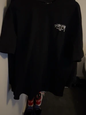 Svart Stüssy t-shirt med tryck - Svart t-shirt från Stüssy med vit logga och text på bröstet samt stort grafiskt tryck med saxofon och städer på ryggen. Klassisk rund hals och korta ärmar. Perfekt för dig som gillar streetwear och vill sticka ut med en ikonisk design. Använd fåtal gånger den är för stor för mig som är 175 cm 