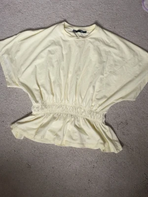 Ljusgul blus med resår i midjan - Säljer en ljusgul blus från Lager 157 i mjuk bomull. Toppen har korta ärmar, rund hals och bred resår i midjan som ger en snygg volang-effekt nedtill. Perfekt för dig som gillar en loose och bekväm passform med lite extra detalj i siluetten. Har använts 1 gång innan.