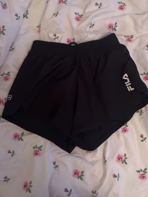 Svarta träningsshorts från FILA XS - Svarta träningsshorts från FILA i storlek XS. De har elastisk midja och är gjorda i lätt polyester som andas. Loggan är tryckt i vitt på ena benet. Använda en eller två gånger