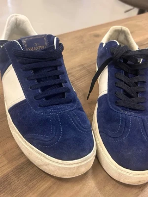 Blåa Valentino Rockrunner sneakers - Snygga blåa och vita Valentino Rockrunner sneakers i mocka med vita detaljer och svart snörning. Klassisk rund tå och platt sula ger en clean look. Perfekta för dig som vill ha något exklusivt och stilrent i garderoben.
