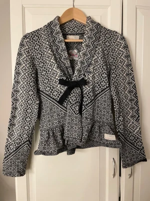 Mönstrad kofta från Odd Molly - Säljer en svartvit kofta från Odd Molly med snyggt geometriskt mönster och dekorativ rosett framtill. Koftan är stickad, har lång ärm och en feminin passform med volangkant nedtill. Perfekt för dig som vill ha något unikt och stilrent!