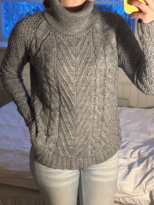 Grå stickad polotröja med kabelmönster - Säljer en grå stickad polotröja med snyggt kabelmönster och bred krage. Tröjan har lång ärm och är ribbad vid muddar och nederkant. Perfekt för kyliga dagar och riktigt mysig att bära. Passar dig som gillar klassisk stil med en modern twist.