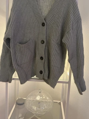 Grå/grön  stickad kofta med knappar - Mysig grå/grön stickad kofta med grov ribbad struktur och stora framfickor. Koftan har v-ringad hals och stängs med fyra svarta knappar framtill. Perfekt för lager-på-lager och chill dagar. Passformen är loose och ärmarna är långärmade.