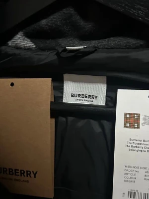 Svart pufferjacka från Burberry - Svart pufferjacka från Burberry med klassisk rutig huva i grått och vitt. Jackan har dragkedja och knappar framtill, två fickor med dragkedja och Burberry-logga på ärmen. Perfekt för kalla dagar och har en riktigt snygg streetstil.
