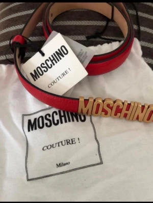 Röd Moschino bälte med gulddetalj - Säljer ett snyggt rött bälte från Moschino med stora guldfärgade bokstäver framtill. Bältet är tillverkat i skinn och har en exklusiv vibe som lyfter vilken outfit som helst. Perfekt för dig som vill sticka ut med en statement-accessoar.
