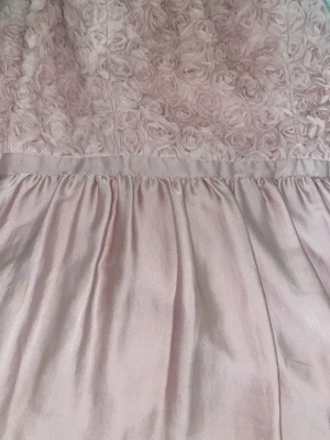 Ljusrosa klänning från Bodyflirt - Supersöt ljusrosa klänning från Bodyflirt i storlek 46. Överdelen har ett romantiskt blommigt 3D-mönster med rosor och kjolen är slät och mjukt glansig. Perfekt för dig som gillar feminina detaljer och vill sticka ut med en unik look.