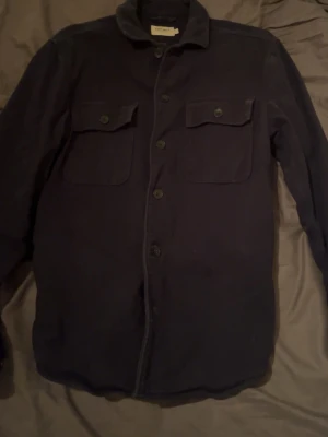 Mörkblå overshirt från East West storlek M - Säljer en stilren mörkblå overshirt från East West med klassisk krage och två stora bröstfickor med knappar. Jackan har knäppning framtill och är långärmad, perfekt för lager-på-lager. Passar dig som gillar enkel och clean look.