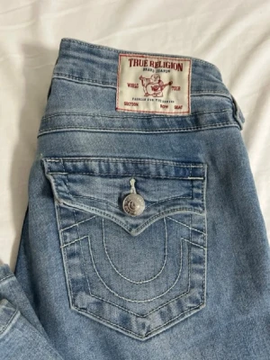 True Religion Joey Low Rise Flare jeans - Säljer ett par ljusblå True Religion Joey Low Rise Flare jeans i storlek 24 då de är förstora. De har snygga broderade fickor med silverknappar och klassisk label bak. Modellen är lågmidjad med bootcut och tillverkad i mjukt jeanstyg. Perfekt för dig som gillar detaljer och ikonisk stil. Byxorna ser lite fläckiga ut men det är min kamera som är trasig då man ser på första bilden att de INTE är fläckiga. Haft i ungefär ett halvår då de är bra skick.