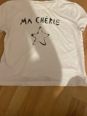 Vit t-shirt från Gina Tricot - Vit t-shirt från Gina Tricot med svart text 'MA CHÉRIE' och en stjärna tryckt både fram och bak. Rund hals och korta ärmar, tillverkad i mjuk bomull. Endast använd fåtal gånger, bra skick!