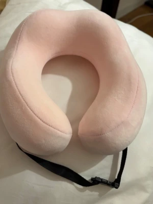 Rosa nackkudde med svart spänne - Supermjuk och skön nackkudde i ljusrosa färg, perfekt för resor. Kudden har ett svart justerbart band med spänne som gör att den sitter på plats. Ytan är len och fluffig, vilket gör den extra bekväm att använda under långa flygturer eller bilresor.