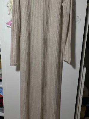 Beige ribbad långklänning - Säljer en beige långklänning med ribbad struktur och långärmade ärmar. Klänningen har en rak och enkel siluett. Materialet känns mjukt och skönt mot huden, och färgen är neutral och lättmatchad. Lite genomskinlig om man har mörka underkläder. 