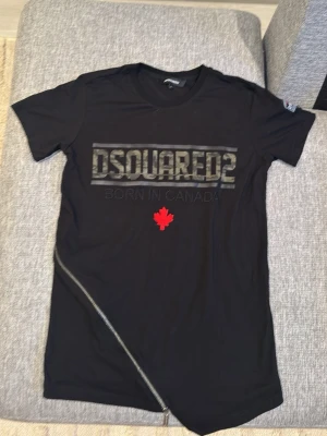 Svart DSQUARED2 t-shirt med dragkedja - Svart t-shirt från DSQUARED2 med camouflagemönstrad logga och texten 'Born in Canada' på bröstet. Röd lönnlöv-detalj under texten och en sned dragkedja nedtill. Kortärmad modell i mjuk bomull, med patch och röd detalj på ena ärmen.