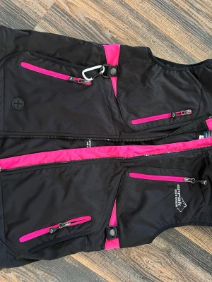 Svart softshellväst från Arrak Outdoor - Säljer en svart softshellväst från Arrak Outdoor med snygga rosa detaljer och dragkedjor. Västen har flera praktiska fickor med dragkedja och en karbinhake framtill. Perfekt för dig som gillar att vara aktiv utomhus