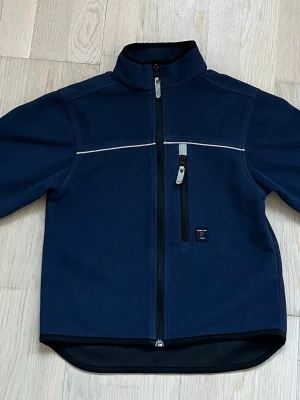 Marinblå fleecejacka Polarn O. Pyret - Säljer en marinblå fleecejacka från Polarn O. Pyret med hel dragkedja, bröstficka med dragkedja och tumhål i ärmsluten. Är storlek 122 och passar barn i 6-8 års åldern. Jackan har en hög krage och reflekterande detaljer både fram och bak. Perfekt för kyliga dagar och enkel att slänga på sig. Kostade originellt ca 300kr.