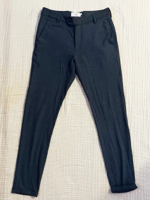 Mörkgrå/blå kostymbyxor Les Deux - Snygga mörkgrå/blå kostymbyxor från Les Deux med diskret fiskbensmönster. Byxorna har smal passform, uppvikta benslut och klassisk midja. Perfekt för dig som gillar stilrena detaljer och vill ha en modern look. Materialet känns mjukt och har en elegant finish.