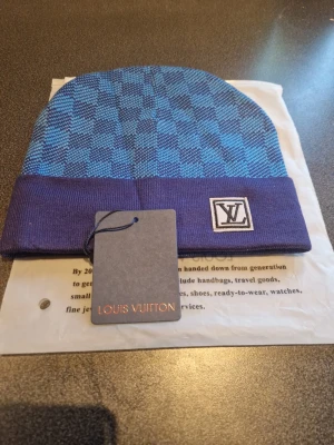 Blå rutig Louis Vuitton mössa - Snygg blå mössa från Louis Vuitton med rutigt mönster och uppvikt kant. Mössan har en vit patch med LV-loggan framtill. Perfekt accessoar för att lyfta din stil under kyliga dagar.