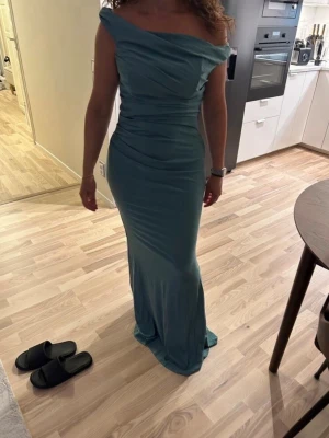 Lång blå offshoulder klänning - Säljer en elegant långklänning i blått med offshoulder-design och figurnära passform. Klänningen har draperade detaljer upptill och är tillverkad i ett mjukt, slätt material som faller snyggt längs kroppen. Perfekt för dig som vill ha en stilren och sofistikerad look.